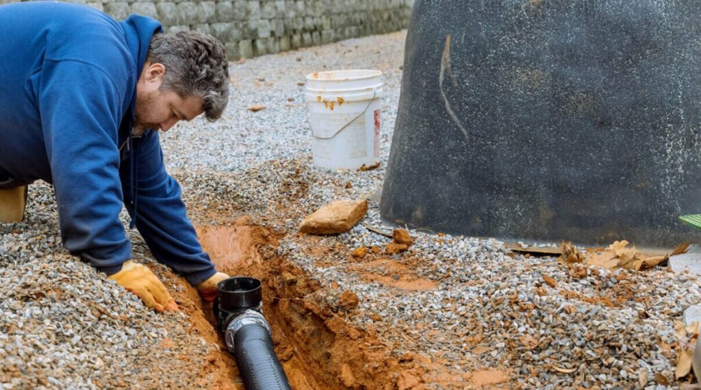 - Happy Home Services | Durham, NC Worker in a blue hoodie kneels beside a trench, connecting a black drainage pipe to a fitting in orange soil and gravel nearby.